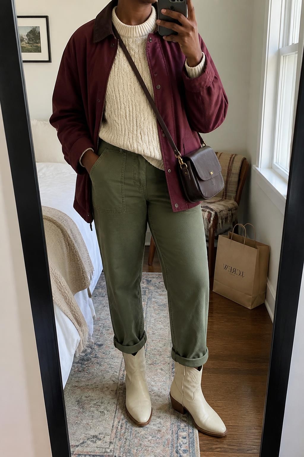 mirror selfie of a Black woman wearing a burgundy cotton barn jacket partly open over an oatmeal cable-knit crewneck sweater, olive straight-leg utility trousers rolled once above cream leather cowboy boots, and a dark brown leather crossbody bag, clearly showing the exact clothing items, colors, fabrics, boots, and bag listed in the Outfit Pieces section, holding a phone in front of a mirror, cropped at NECK so the face does not show, relaxed outdoor-ready selfie pose in a cozy bedroom mirror setup, natural window light, with a folded blanket on a chair and shopping bags beside it, realistic cotton, cable knit, utility fabric, and leather textures, latest iPhone photo quality