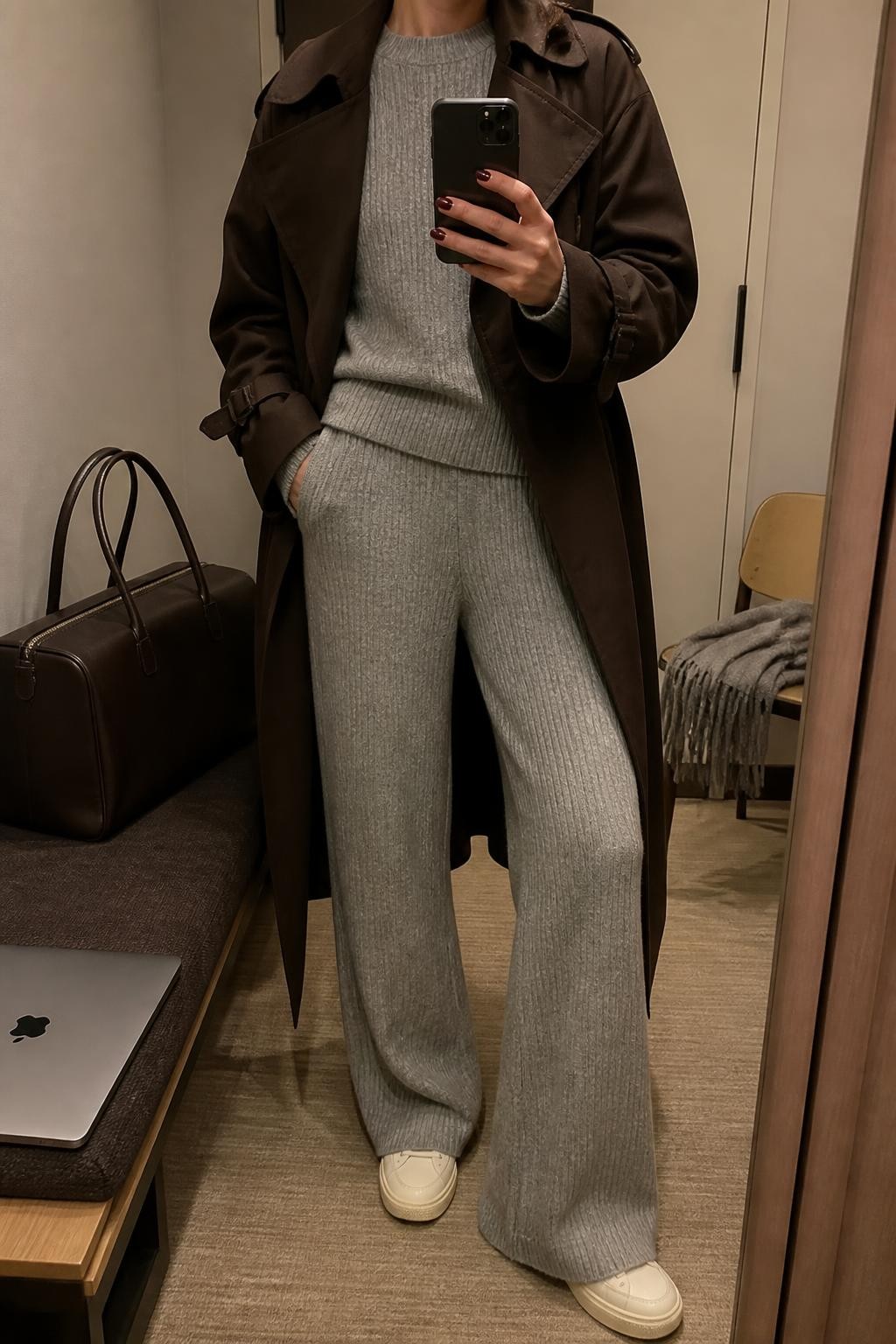 mirror selfie of a woman wearing an open chocolate brown cotton-blend trench coat over a matching heather gray ribbed crewneck sweater and heather gray ribbed wide-leg knit trousers, cream leather sneakers, and a dark brown structured travel tote with a top zip, clearly showing the exact clothing items, colors, fabrics, sneakers, and bag listed in the Outfit Pieces section, holding a phone in front of a mirror, cropped at NECK so the face does not show, relaxed travel-ready selfie pose in a minimal dressing room, warm moody lighting, with a laptop resting on a bench and a folded blanket on a chair, realistic cotton, ribbed knit, leather, and structured bag textures, latest iPhone photo quality
