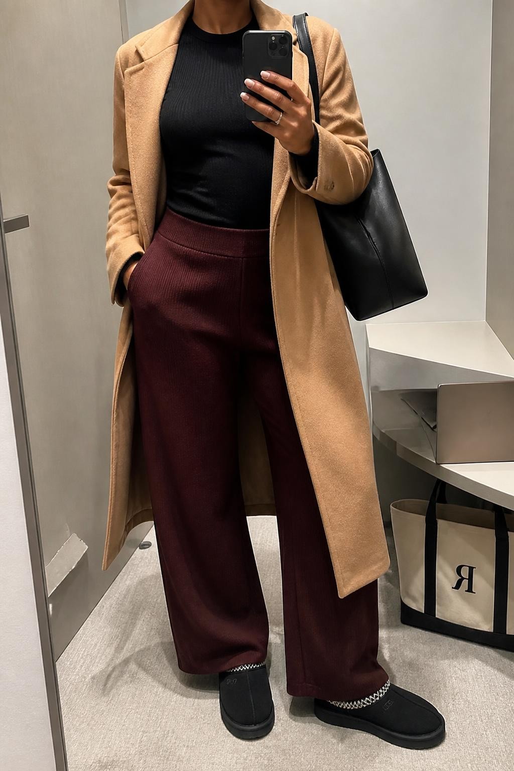 mirror selfie of a Black woman wearing an open camel longline wool-blend coat over a black fitted ribbed long-sleeve top tucked into burgundy wide-leg knit trousers, black Tasman Ugg slippers, and a black structured travel tote, clearly showing the exact clothing items, colors, fabrics, slippers, and bag listed in the Outfit Pieces section, holding a phone in front of a mirror, cropped at NECK so the face does not show, relaxed travel-ready selfie pose in a minimal dressing room, soft indoor lighting, with a laptop resting on a bench and a tote bag on the floor nearby, realistic wool, ribbed knit, soft trouser knit, suede, and structured bag textures, latest iPhone photo quality