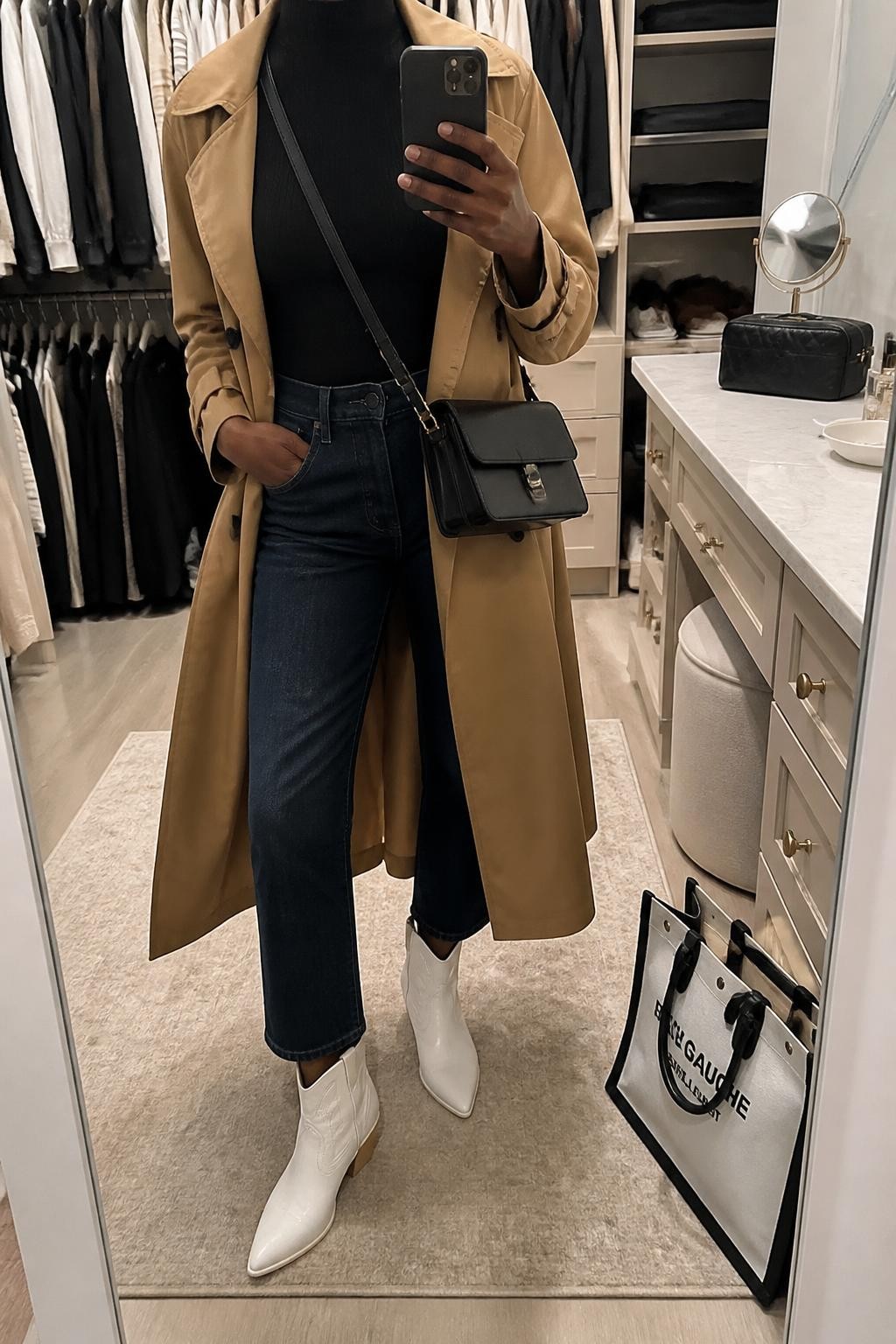 mirror selfie of a Black woman wearing an open camel belted cotton trench coat over a black fitted ribbed mock-neck top, dark-wash cropped straight-leg jeans stopping above white leather cowboy boots, and a black structured crossbody bag, clearly showing the exact clothing items, colors, fabrics, boots, and bag listed in the Outfit Pieces section, holding a phone in front of a mirror, cropped at NECK so the face does not show, relaxed travel-ready selfie pose in a walk-in closet mirror, soft indoor lighting, with a tote bag resting on the floor and a cosmetic bag on the vanity, realistic cotton, ribbed knit, denim, and leather textures, natural unstaged mirror photo, latest iPhone photo quality