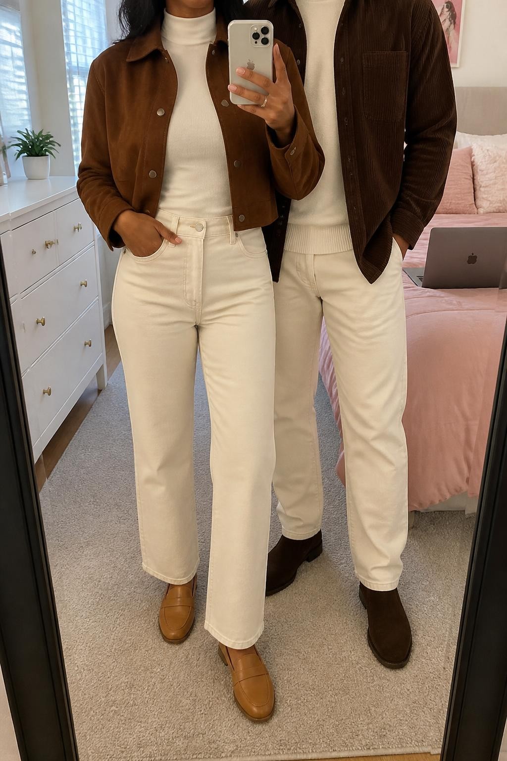 mirror selfie of a Black couple wearing coordinated chocolate brown and ecru fall outfits, the woman wearing an open chocolate cropped suede jacket over an ivory fitted mock-neck top, ecru straight-leg jeans, and tan leather loafers, the man wearing an open chocolate corduroy overshirt over an ivory crewneck sweater, ecru jeans, and dark brown suede boots, clearly showing the exact clothing items, colors, fabrics, and footwear listed in the Outfit Pieces section, the woman holding a phone while the man stands beside her, both cropped at NECK so their faces do not show, relaxed polished couple pose in a feminine-themed bedroom, natural window light, with a small plant on a dresser and a laptop on the bed, realistic suede, corduroy, knit, denim, and leather textures, latest iPhone photo quality
