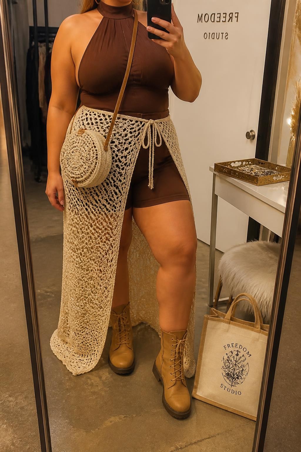 A realistic fashion mirror selfie of a plus-size adult woman wearing a refined bohemian festival outfit: a cream open-crochet maxi skirt styled as a layered fashion piece over fitted chocolate-brown high-waisted shorts, paired with a matching chocolate-brown halter-neck top featuring a supportive wide neck band. She wears tan lace-up boots and carries a round cream woven crossbody bag. She is holding a smartphone in front of a full-length mirror, with the composition cropped below the chin so her face is not visible. Relaxed, confident bohemian fashion pose in a stylish fashion-studio mirror corner with warm evening golden-hour lighting, a tote bag resting on the floor and a jewelry tray on the vanity. Refined contemporary boho festival styling, realistic crochet and woven textures, coordinated earthy colors, natural proportions, and high-quality modern smartphone photography.