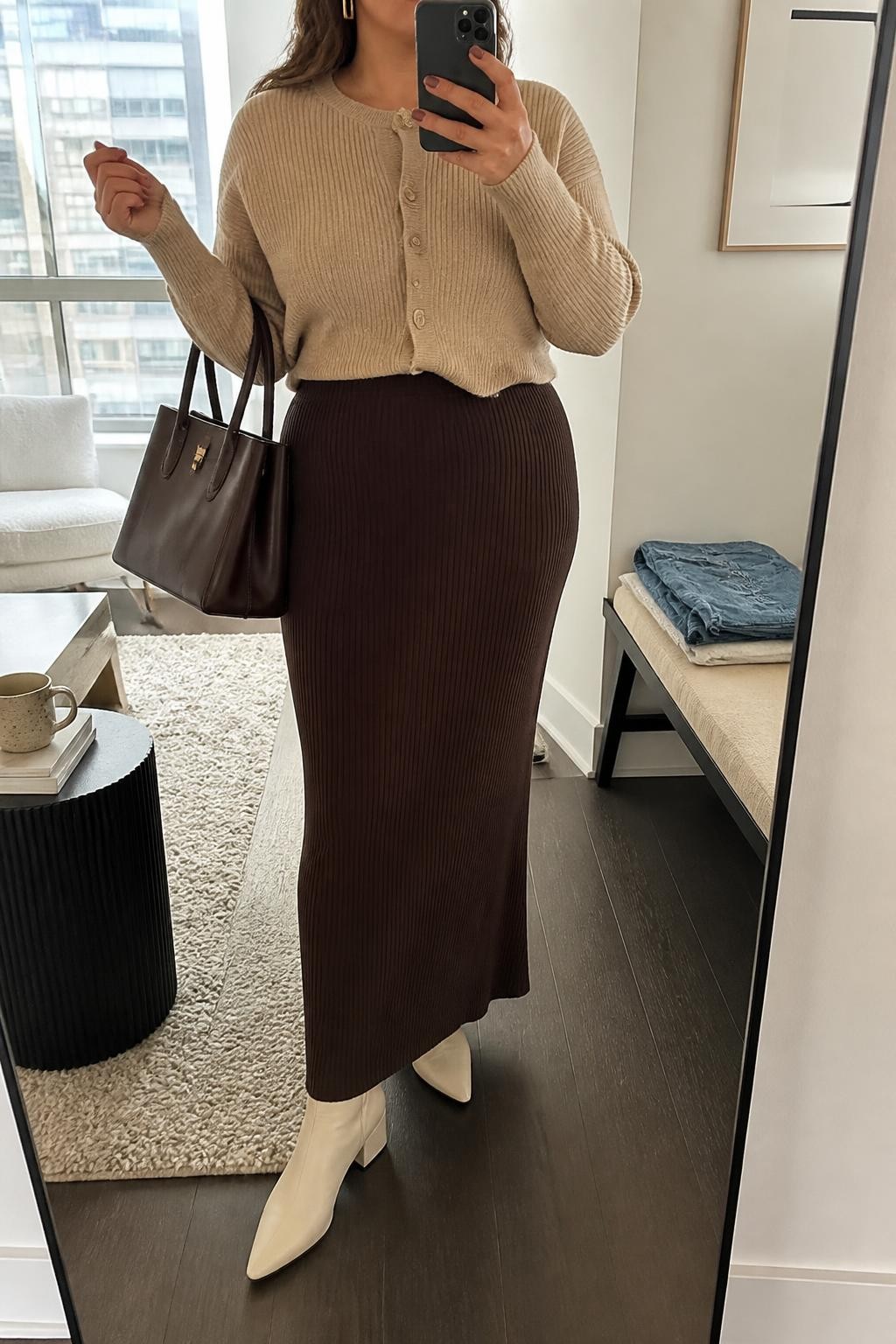 Mirror selfie of a plus size woman wearing a chocolate brown ribbed knit high-waisted maxi skirt with a softly fitted shape, a beige cropped cardigan buttoned at the front, cream pointed ankle boots with low block heels, a structured chocolate brown handbag, and gold rectangular earrings, clearly showing the exact clothing items, chocolate knit, beige cardigan, colors, footwear, and accessories listed, holding a phone in front of a mirror, cropped at the neck so her face is hidden, relaxed polished selfie pose, in a stylish city apartment with soft indoor lighting, a coffee mug on a side table and folded denim jeans on a bench, realistic autumn outfit, latest iPhone photo quality.