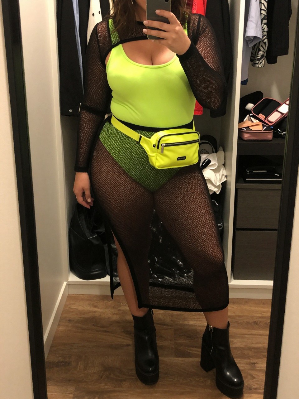 Mirror selfie of a plus size woman wearing a black mesh long-sleeve midi dress with a side slit over a fitted neon lime scoop-neck bodysuit, black platform ankle boots, and a compact neon lime belt bag, clearly showing the exact clothing layers, black mesh, neon lime bodysuit, side slit, footwear, and accessories listed, holding a phone in front of a mirror, cropped at the neck so the face is hidden, relaxed daring selfie pose, in a walk-in closet mirror with warm moody lighting, an open closet with clothes visible and a cosmetic bag on the counter, realistic late-night festival outfit, latest iPhone photo quality.