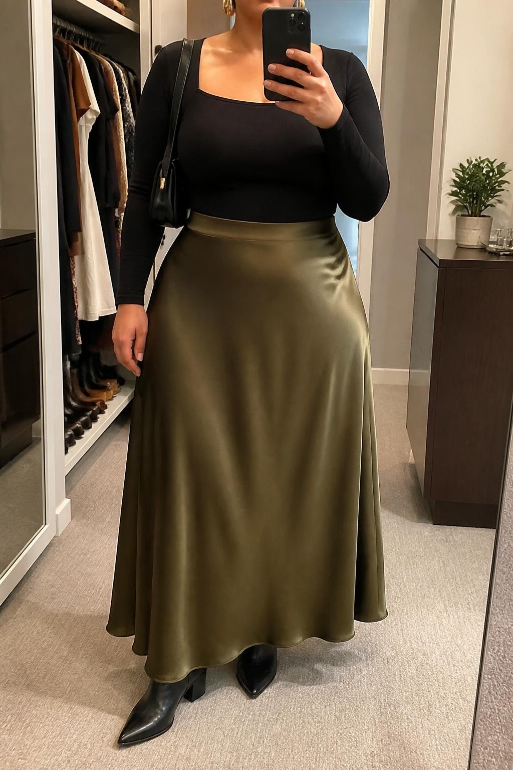 Mirror selfie of a plus size woman wearing an olive green high-waisted satin maxi skirt with a fluid A-line shape, a fitted black long-sleeve square-neck top, black pointed ankle boots with low block heels, a compact black leather shoulder bag, and chunky gold cuff earrings, clearly showing the exact clothing items, olive satin, black fitted top, footwear, and accessories listed, holding a phone in front of a mirror, cropped at the neck so her face does not appear, relaxed confident selfie stance, in a walk-in closet mirror with soft indoor lighting, an open closet with clothes visible and a small plant on a dresser, realistic autumn dinner outfit, latest iPhone photo quality.