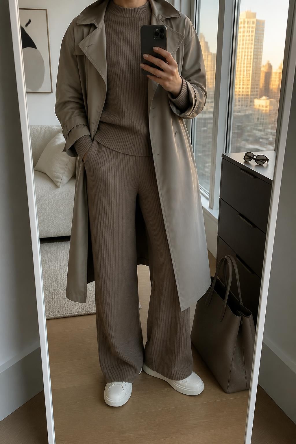 mirror selfie of a woman wearing an open taupe water-resistant cotton trench coat over a matching mushroom brown ribbed crewneck sweater and mushroom brown ribbed wide-leg knit trousers, cream leather sneakers, and a dark taupe structured travel tote, clearly showing the exact clothing items, colors, fabrics, sneakers, and bag listed in the Outfit Pieces section, holding a phone in front of a mirror, cropped at NECK so the face does not show, relaxed travel-ready selfie pose in a stylish city apartment, evening golden hour light, with a tote bag resting on the floor and a pair of sunglasses on a dresser, realistic cotton, ribbed knit, leather, and structured bag textures, latest iPhone photo quality
