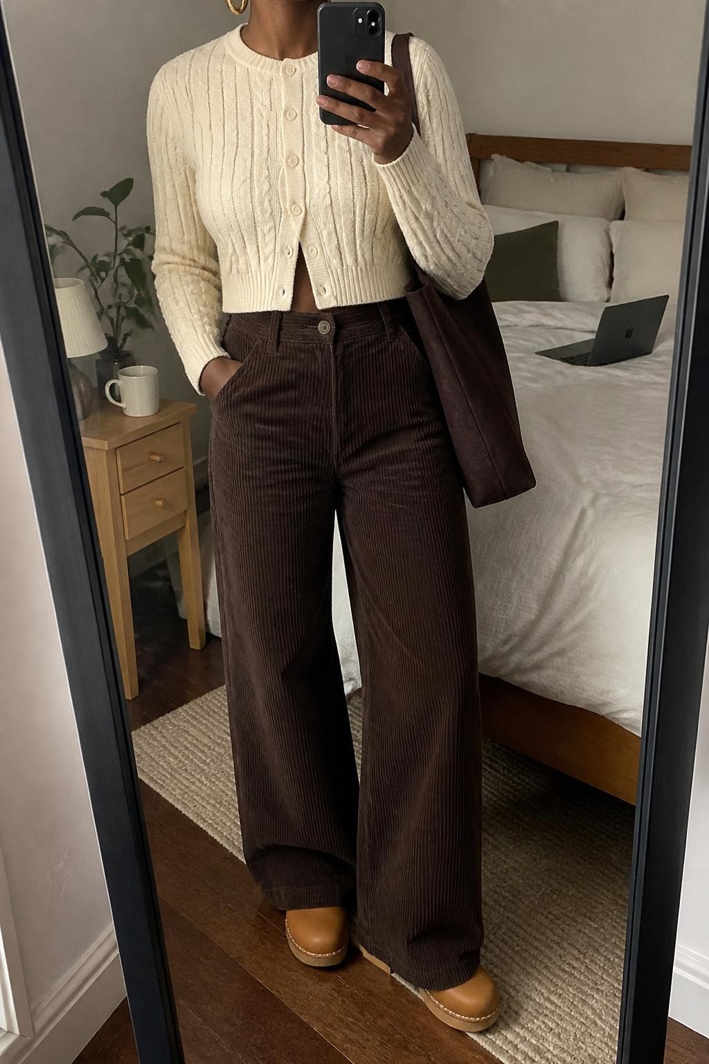 Mirror selfie of a Black woman wearing a cropped cream cable-knit cardigan, high-waisted chocolate-brown wide-leg corduroy pants, tan leather clogs, a dark brown canvas tote bag, and small hammered-gold hoop earrings, clearly showing the exact clothing items, colors, fabrics, and accessories listed, holding a phone in front of a cozy bedroom mirror setup, cropped at the neck so the face does not show, relaxed casual selfie pose, soft indoor lighting, a coffee mug on a bedside table and a laptop on the bed, natural lived-in environment, latest iPhone photo quality.