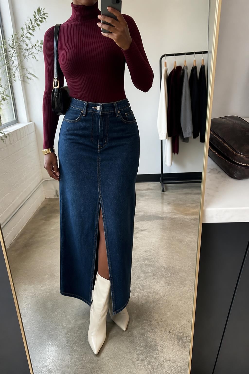 mirror selfie of a Black woman wearing a burgundy fitted mock-neck knit sweater tucked smoothly into a dark-wash front-slit denim maxi skirt with the slit centered over cream pointed-toe ankle boots, a black structured shoulder bag, and a brushed gold cuff bracelet, clearly showing the exact clothing items, colors, fabrics, boots, bag, and bracelet listed in the Outfit Pieces section, holding a phone in front of a mirror, cropped at NECK so the face does not show, relaxed confident selfie pose in a fashion studio mirror corner, natural window light, with a clothing rack holding a few garments behind her and a cosmetic bag on a counter, realistic knit, denim, leather-look, and metal textures, latest iPhone photo quality