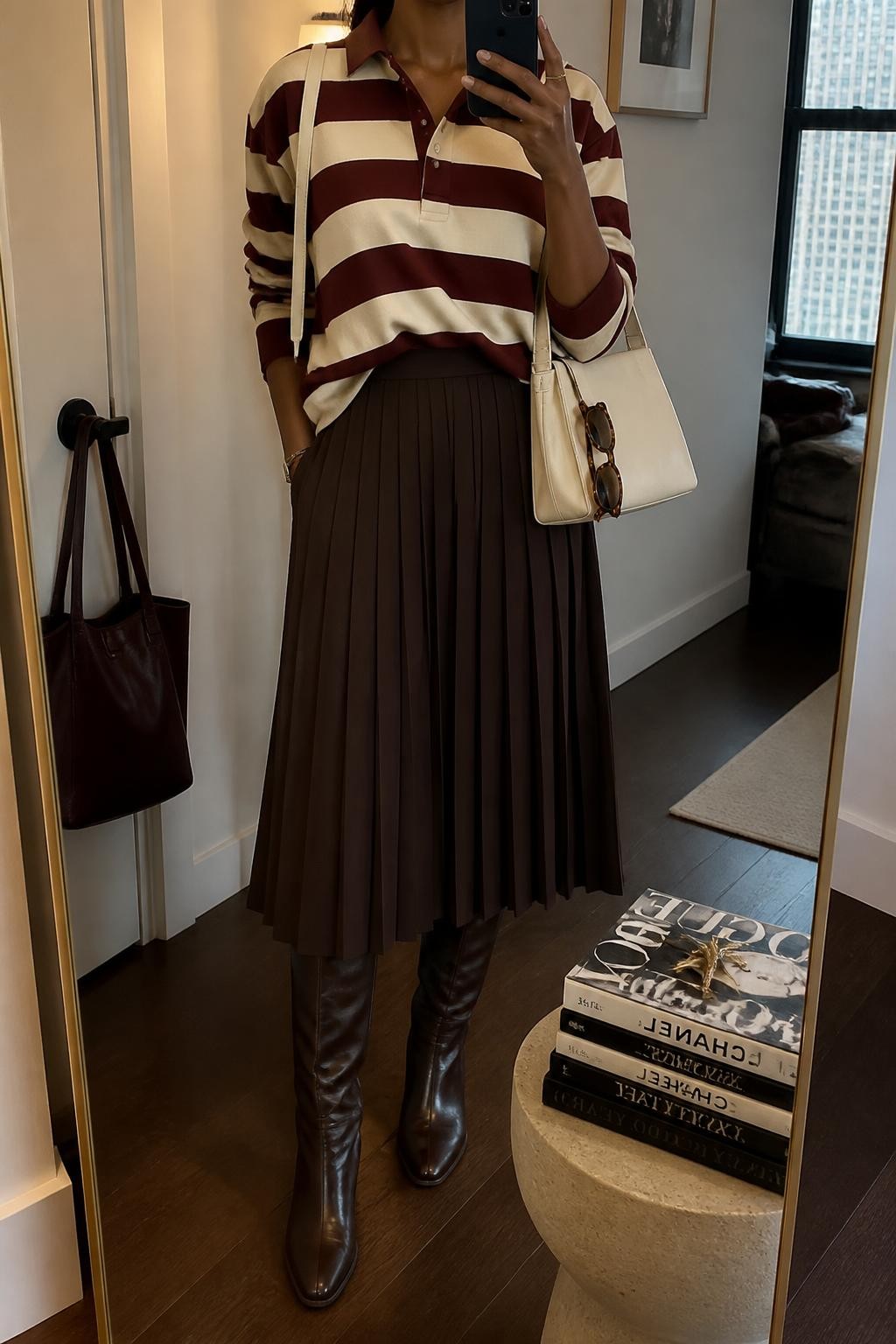 mirror selfie of a Black woman wearing a burgundy and cream striped rugby shirt with a small front tuck into a chocolate brown pleated midi skirt, dark brown leather-look knee-high boots, a cream structured shoulder bag, and tortoiseshell rectangular sunglasses hooked onto the bag, clearly showing the exact clothing items, colors, striped pattern, fabrics, boots, bag, and sunglasses listed in the Outfit Pieces section, holding a phone in front of a mirror, cropped at NECK so the face does not show, relaxed preppy selfie pose in a stylish city apartment, warm moody lighting, with a stack of fashion magazines on a side table and a handbag hanging on a door handle, realistic cotton-look fabric, pleated material, leather-look, and tortoiseshell textures, latest iPhone photo quality