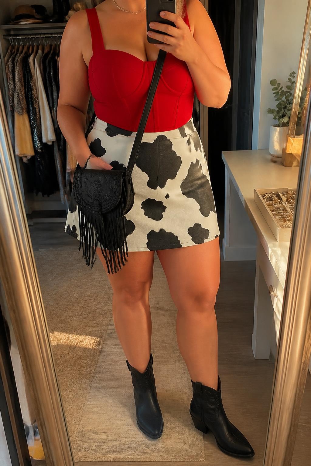 A realistic contemporary fashion mirror selfie featuring an adult plus-size woman wearing a Western-inspired festival outfit. She wears a black-and-white cow-print A-line skirt with a structured red fashion top featuring wide supportive straps, paired with black Western ankle boots. A black leather fringe crossbody bag and slim silver chain necklace complete the coordinated look. She holds a smartphone in front of a full-length mirror, with the framing cropped below the chin so her face is outside the composition. Relaxed, confident fashion pose in a stylish walk-in closet with warm evening golden-hour lighting, an open wardrobe with clothing visible in the background, and a jewelry tray on the vanity. Polished Western festival fashion photography, realistic textile and leather textures, crisp cow-print pattern, detailed fringe, coordinated colors, natural body proportions, authentic indoor lighting, and high-quality modern smartphone photography.