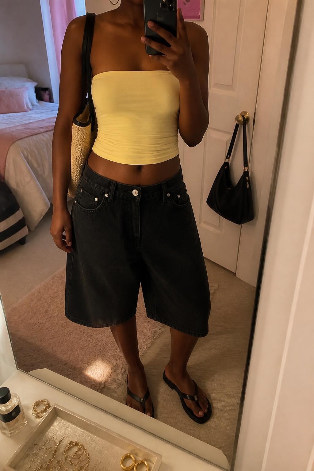 mirror selfie of a Black woman wearing loose mid-rise baggy black denim jorts, a fitted butter yellow stretch cotton tube top, slim black flat sandals, a natural woven shoulder bag with black trim and medium gold hoop earrings, wearing exactly the listed outfit with no extra clothing pieces, holding a phone in front of a mirror, cropped at NECK so the face does not show, relaxed casual selfie pose, in a feminine-themed bedroom, evening golden hour, jewelry tray on the vanity and a handbag hanging on a door handle, realistic casual styling, latest iPhone photo quality.
