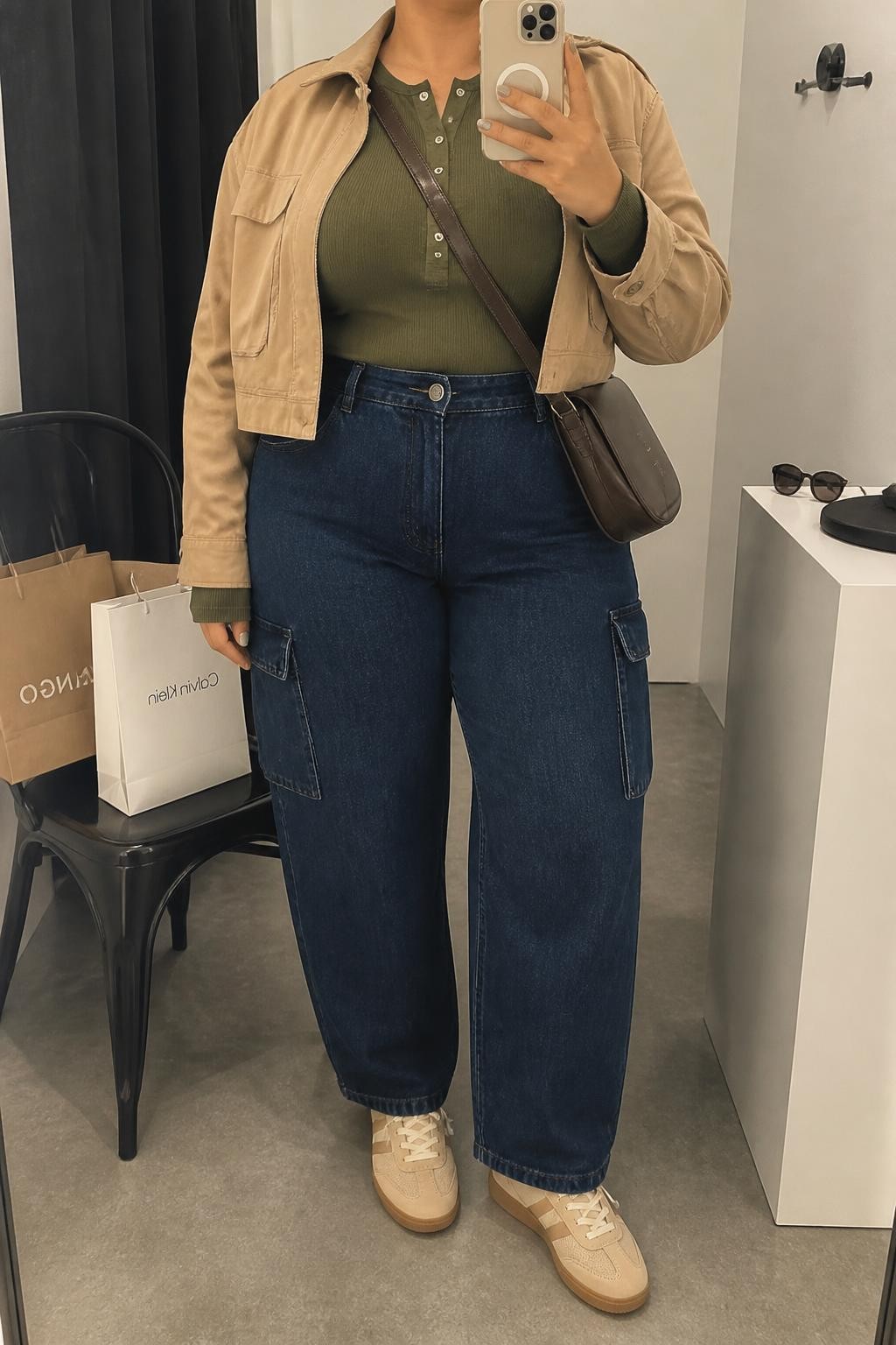 Mirror selfie of a plus size woman wearing indigo high-rise cargo jeans with flat side pockets, a fitted olive green ribbed Henley top, a cropped tan utility jacket, a dark brown crossbody bag, and beige lace-up sneakers, clearly showing the exact clothing pieces, colors, textures, pockets, and accessories listed, holding a phone in front of a mirror, cropped at the neck so the face does not show, relaxed casual selfie pose, in a fashion studio mirror corner with soft indoor lighting, shopping bags beside a chair and sunglasses placed on a counter, realistic modern iPhone photograph, latest iPhone photo quality.