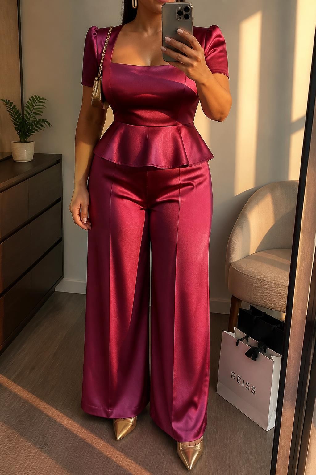 Mirror selfie of a plus size woman wearing a berry pink satin peplum top with a square neckline and short structured sleeves, matching berry satin high-waisted palazzo trousers, metallic gold pointed pumps, a small gold chain-strap bag, and gold rectangular earrings, clearly showing the exact coordinated outfit, berry satin, peplum shape, trousers, footwear, and accessories listed, holding a phone in front of a mirror, cropped at the neck so the face is hidden, relaxed confident selfie stance, in a minimal dressing room with evening golden-hour lighting, shopping bags beside a chair and a small plant on a dresser, realistic fashion-forward wedding guest appearance, latest iPhone photo quality.
