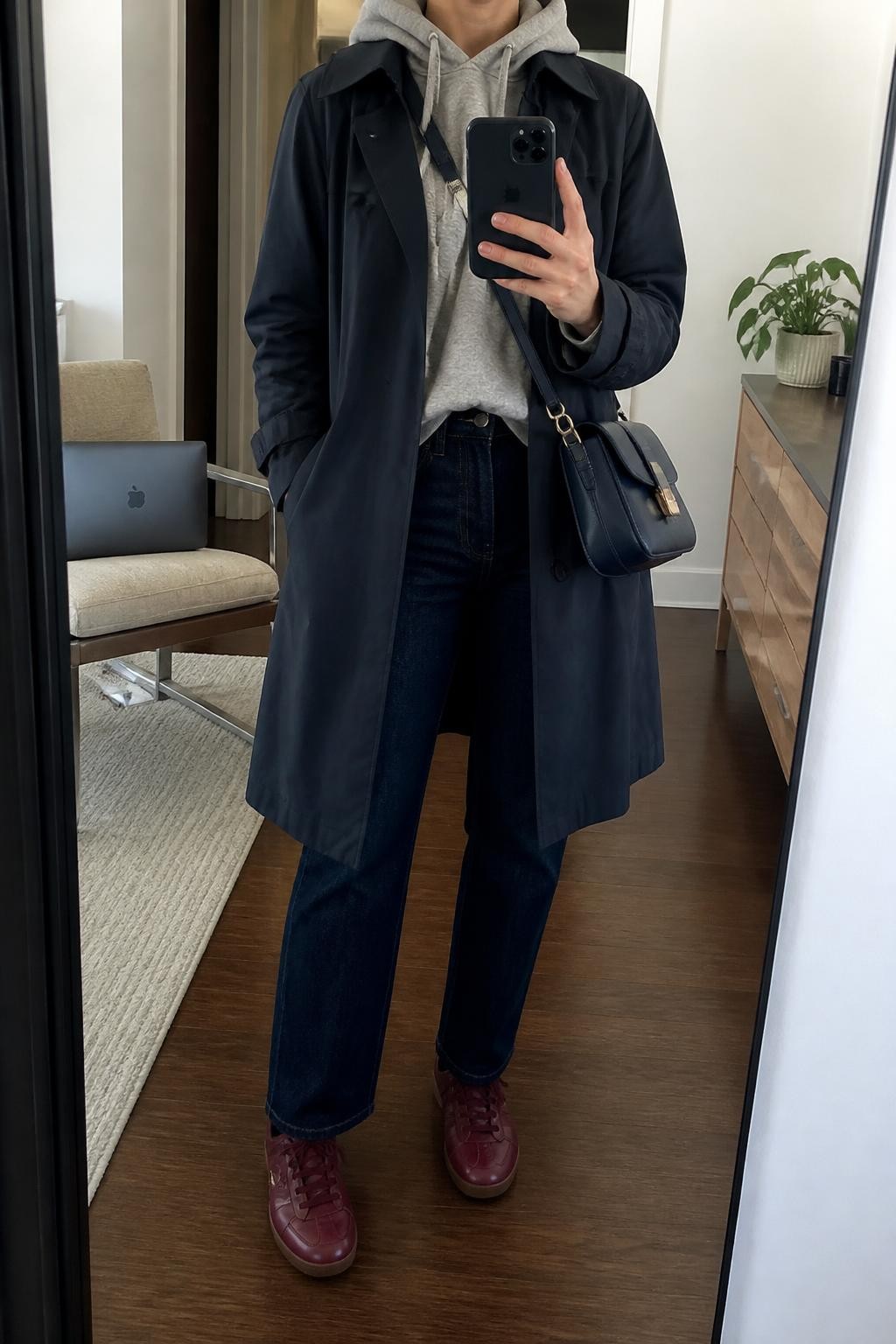 mirror selfie of a woman wearing an open navy water-resistant cotton trench coat over a heather gray brushed-cotton hoodie with the hood placed neatly outside the coat collar, dark indigo straight-leg jeans, burgundy leather sneakers, and a navy structured crossbody bag, clearly showing the exact clothing items, colors, fabrics, sneakers, and bag listed in the Outfit Pieces section, holding a phone in front of a mirror, cropped at NECK so the face does not show, relaxed casual selfie pose in a stylish city apartment, soft indoor lighting, with a laptop resting on a chair and a small plant on a dresser, realistic cotton, fleece, denim, and leather textures, latest iPhone photo quality