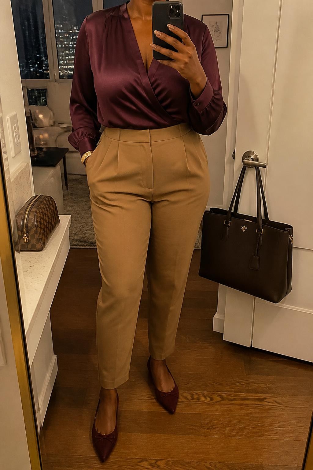 mirror selfie of a Black curvy woman wearing a plum satin wrap-front blouse secured comfortably with the lower front edge tucked into camel high-waisted tapered trousers, burgundy pointed-toe ballet flats, a dark brown structured handbag, and a slim gold cuff bracelet, clearly showing the exact clothing items, colors, fabrics, shoes, bag, and bracelet listed in the Outfit Pieces section, holding a phone in front of a mirror, cropped at NECK so the face does not show, relaxed professional selfie pose in a stylish city apartment, warm moody lighting, with a handbag hanging on a door handle and a cosmetic bag on a nearby counter, realistic satin, trouser, and leather textures, latest iPhone photo quality