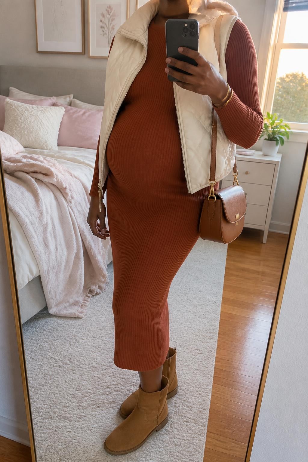 mirror selfie of a Black pregnant woman wearing a rust ribbed maternity sweater dress fitted comfortably around her visible baby bump, an open cream cropped quilted vest, tan suede flat ankle boots, a cognac leather saddle bag, and a slim gold cuff bracelet, clearly showing the exact clothing items, colors, fabrics, boots, bag, and bracelet listed in the Outfit Pieces section, holding a phone in front of a mirror, cropped at NECK so the face does not show, relaxed maternity selfie pose in a feminine-themed bedroom, evening golden hour light, with soft pillows on an unmade bed and a small plant on a dresser, realistic ribbed knit, quilting, suede, and leather textures, natural pregnancy proportions, latest iPhone photo quality