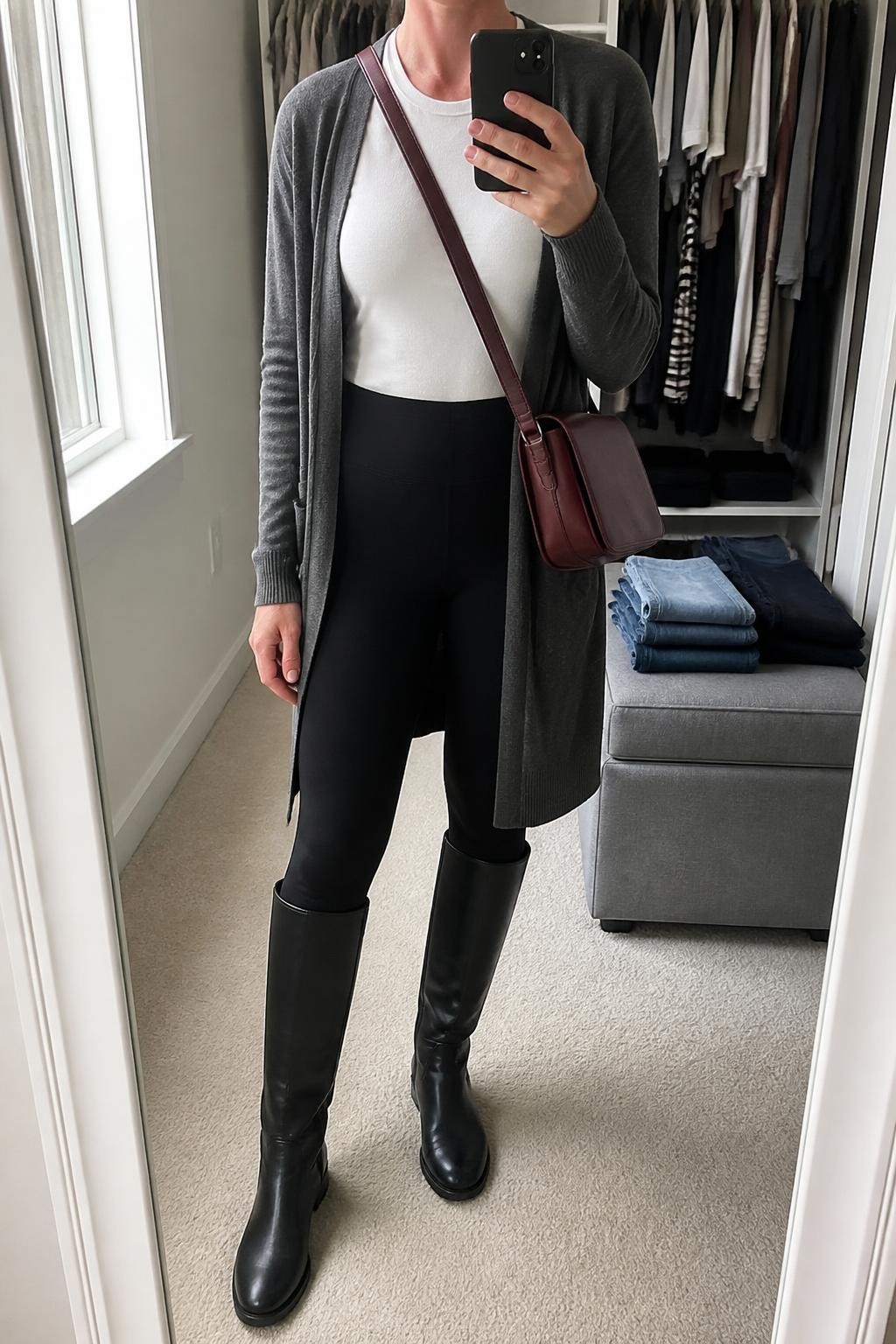 Mirror selfie of a woman wearing a long charcoal-grey open-front cardigan, a fitted white crew-neck T-shirt, matte black high-waisted leggings, black leather riding boots, and a structured oxblood crossbody bag, clearly showing the exact clothing items, colors, fabrics, and accessories listed, holding a phone in front of a walk-in closet mirror, cropped at the neck so the face does not show, relaxed casual selfie pose, natural window light, an open closet with clothes visible and folded denim jeans on a bench, casual and uncluttered, latest iPhone photo quality.