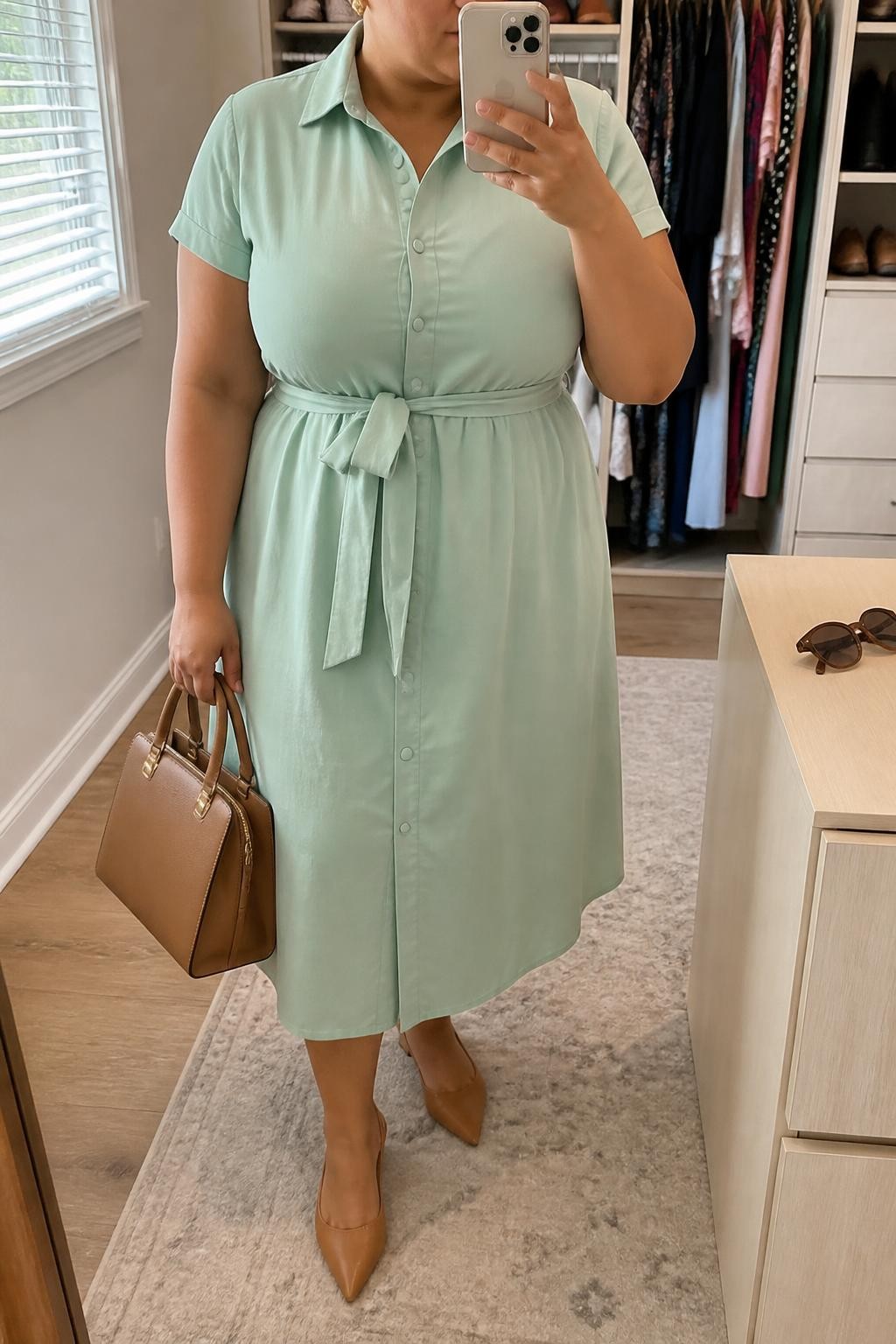 Mirror selfie of a plus size woman wearing a mint green cotton shirt midi dress with short sleeves, a button front, and a matching fabric belt, tan pointed slingback flats, a structured tan leather handbag, and small gold stud earrings, clearly showing the exact dress, mint cotton, fabric belt, tan footwear, and accessories listed, holding a phone in front of a mirror, cropped at the neck so her face does not appear, relaxed neat selfie stance, in a walk-in closet mirror with natural window light, an open closet with clothes visible and sunglasses placed on a dresser, realistic casual baby shower outfit, latest iPhone photo quality.