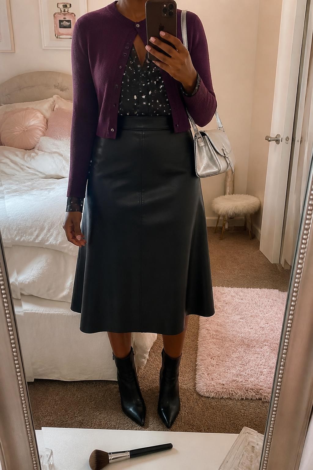 mirror selfie of a Black woman wearing a plum fine-knit cropped cardigan buttoned only at the top over a black chiffon blouse with a clear small silver star print tucked into a black A-line faux-leather midi skirt, black pointed-toe ankle boots, and a silver metallic shoulder bag, clearly showing the exact clothing items, colors, star print, fabrics, boots, and bag listed in the Outfit Pieces section, holding a phone in front of a mirror, cropped at NECK so the face does not show, relaxed polished selfie pose in a feminine-themed bedroom, soft indoor lighting, with a makeup brush on the counter and soft pillows on an unmade bed, realistic knit, chiffon, faux-leather, leather, and metallic textures, latest iPhone photo quality