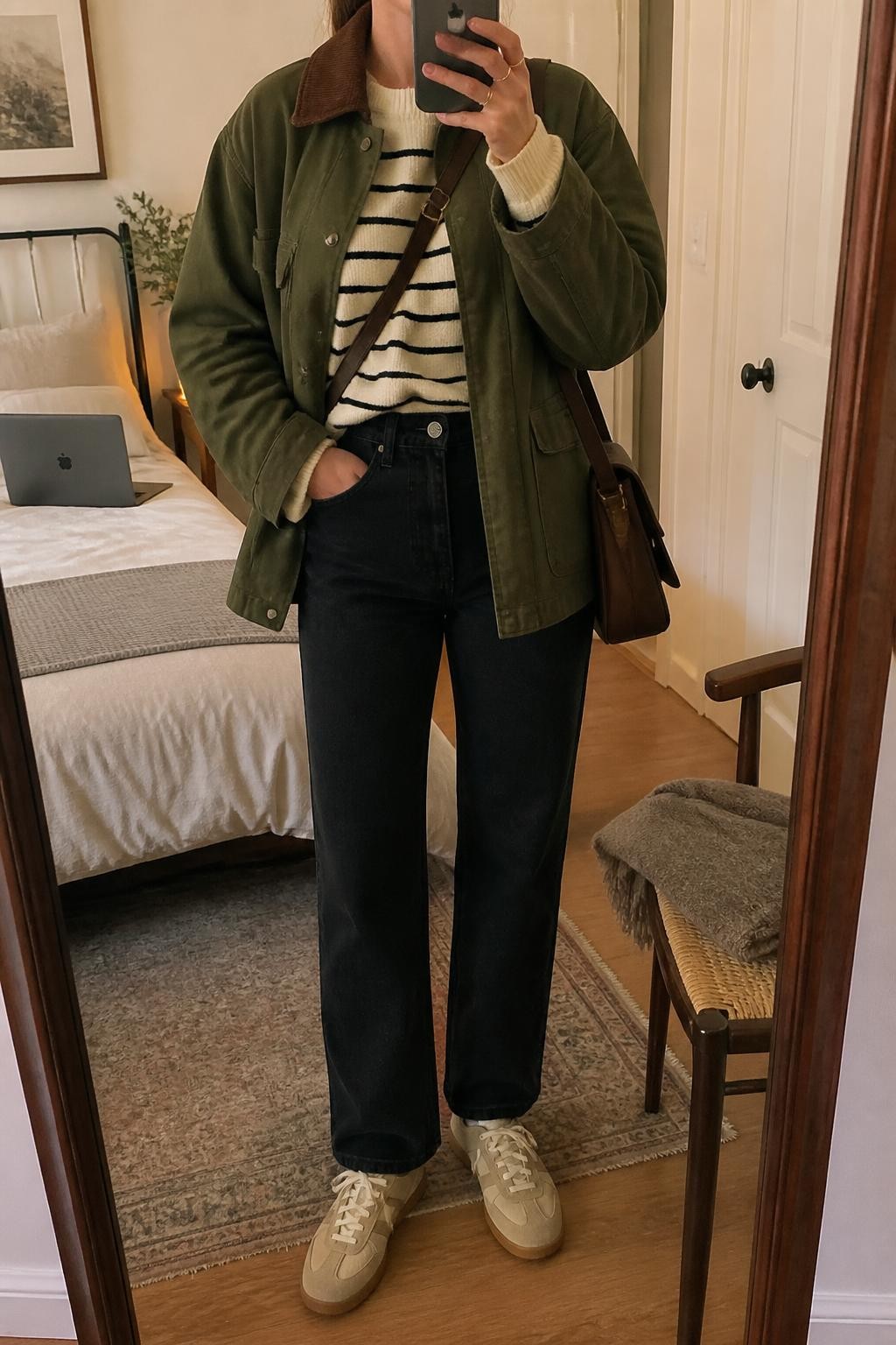 mirror selfie of a woman wearing an open olive cotton barn jacket with a brown corduroy collar over a cream and black striped knit sweater, black high-rise straight-leg jeans, beige retro-style suede sneakers, and a chocolate canvas crossbody bag, clearly showing the exact clothing items, colors, striped pattern, fabrics, sneakers, and bag listed in the Outfit Pieces section, holding a phone in front of a mirror, cropped at NECK so the face does not show, relaxed casual selfie pose in a cozy bedroom mirror setup, warm moody lighting, with a folded blanket on a chair and a laptop on the bed, realistic cotton, corduroy, knit, denim, suede, and canvas textures, latest iPhone photo quality
