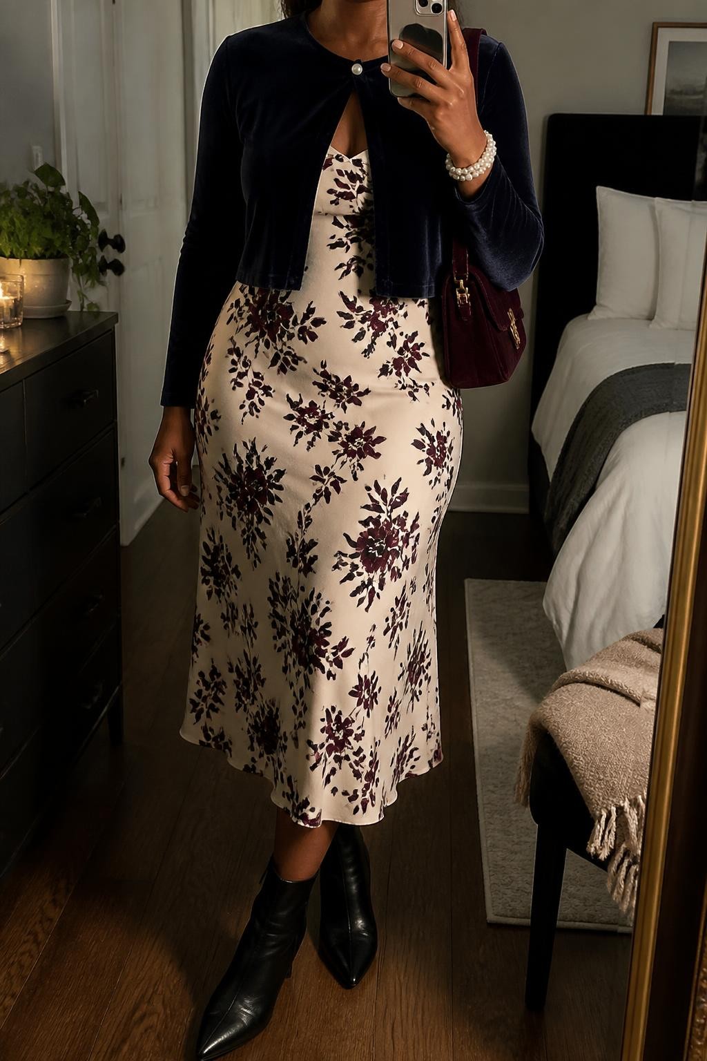 Image Prompt mirror selfie of a Black woman wearing a navy velvet cropped cardigan with pearl buttons fastened only at the top over a cream satin midi slip dress with a clear burgundy floral print, black leather ankle boots, a burgundy velvet mini shoulder bag, and a pearl cuff bracelet, clearly showing the exact clothing items, colors, floral print, fabrics, buttons, boots, bag, and bracelet listed in the Outfit Pieces section, holding a phone in front of a mirror, cropped at NECK so the face does not show, relaxed elegant selfie pose in a cozy bedroom mirror setup, warm moody lighting, with a small plant on a dresser and a folded blanket on a chair, realistic velvet, pearl, satin, printed fabric, and leather textures, latest iPhone photo quality
