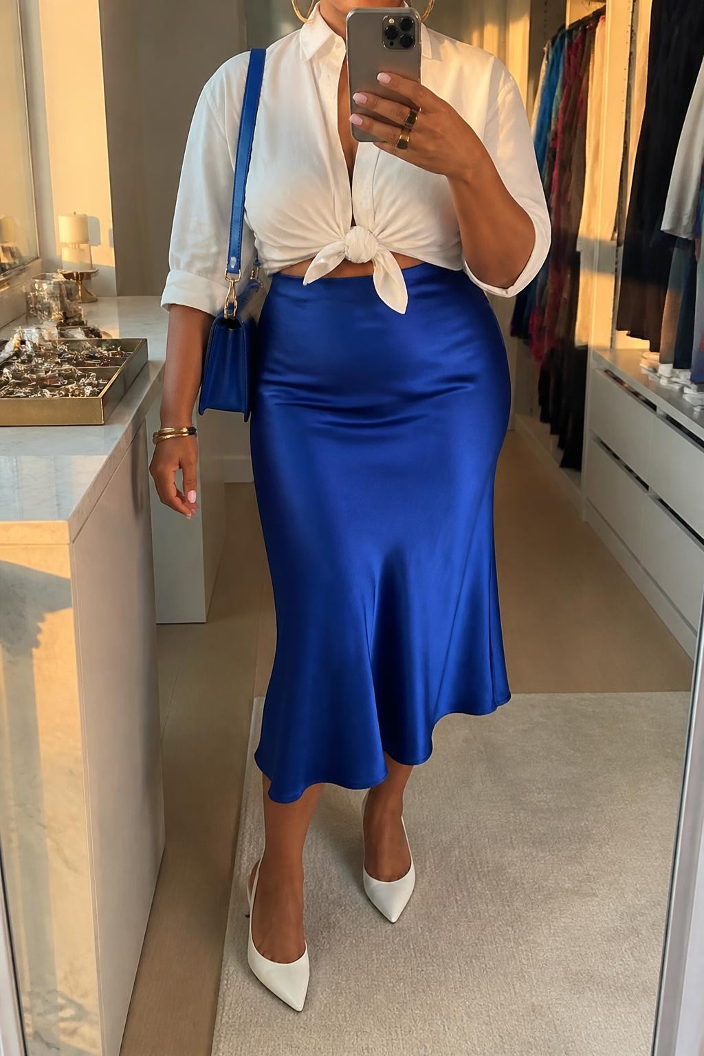 Mirror selfie of a plus size woman wearing a cobalt blue high-waisted satin midi skirt with a fluid bias-cut shape, a white cotton cropped button-down shirt tied softly at the waist, white pointed kitten heels, a structured cobalt shoulder bag, and silver hoop earrings, clearly showing the exact clothing items, cobalt satin, white cotton shirt, colors, footwear, and accessories listed, holding a phone in front of a mirror, cropped at the neck so her face does not appear, relaxed bright selfie stance, in a walk-in closet mirror with evening golden-hour lighting, an open closet with clothes visible and a jewelry tray on the vanity, realistic summer dinner outfit, latest iPhone photo quality.