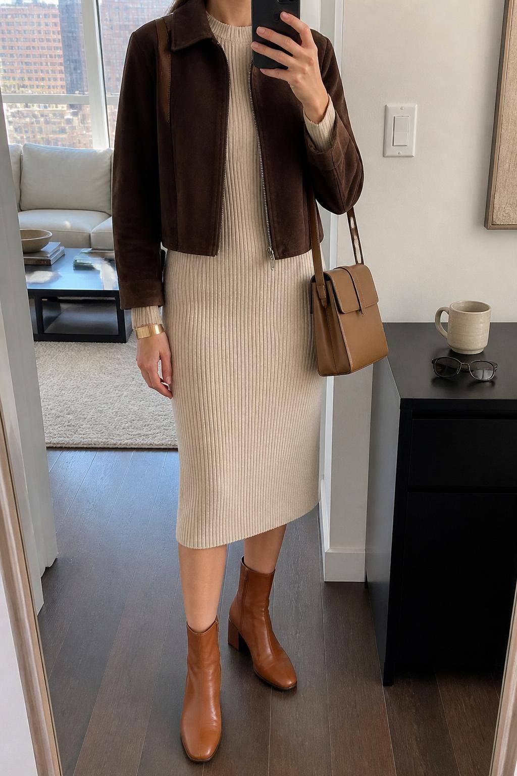 mirror selfie of a woman wearing an open chocolate brown cropped suede jacket over an oatmeal ribbed long-sleeve midi dress with the hem stopping slightly above cognac leather block-heel ankle boots, a tan structured shoulder bag, and a brushed gold cuff bracelet, clearly showing the exact clothing items, colors, fabrics, boots, bag, and bracelet listed in the Outfit Pieces section, holding a phone in front of a full-length mirror, cropped at NECK so the face does not show, relaxed polished selfie pose in a stylish city apartment, natural window light, with a coffee mug on a side table and sunglasses placed on a dresser, realistic suede, ribbed knit, and leather textures, natural unstaged composition, latest iPhone photo quality
