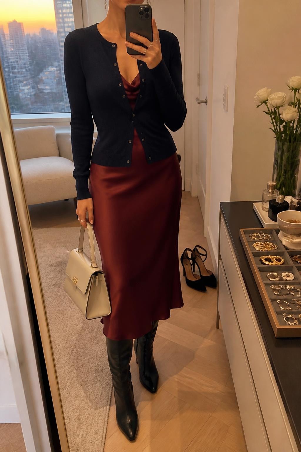 Mirror selfie of a woman wearing a fitted navy cardigan layered over a burgundy satin midi slip dress, black leather knee-high boots, a structured cream handbag, and delicate silver drop earrings, clearly showing the exact clothing items, colors, fabrics, and accessories listed, holding a phone in front of a stylish city apartment mirror, cropped at the neck so the face does not show, relaxed casual selfie pose, evening golden-hour lighting, a jewelry tray on the vanity and a pair of heels on the floor nearby, elegant natural atmosphere, latest iPhone photo quality.