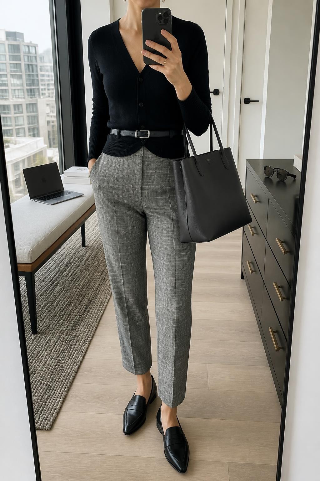 Mirror selfie of a woman wearing tailored grey plaid ankle-length trousers, a fitted black V-neck cardigan fastened and belted at the waist, black leather pointed-toe loafers, a structured charcoal tote bag, and delicate silver stud earrings, clearly showing the exact clothing items, colors, fabrics, and accessories listed, holding a phone in front of a stylish city apartment mirror, cropped at the neck so the face does not show, relaxed confident selfie pose, natural window light, a laptop on a bench and sunglasses placed on a dresser, realistic office-ready setting, latest iPhone photo quality.
