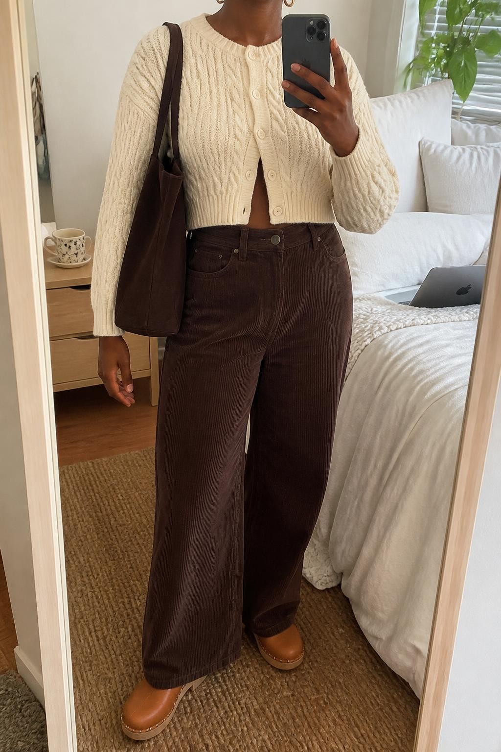 Mirror selfie of a Black woman wearing a cropped cream cable-knit cardigan, high-waisted chocolate-brown wide-leg corduroy pants, tan leather clogs, a dark brown canvas tote bag, and small hammered-gold hoop earrings, clearly showing the exact clothing items, colors, fabrics, and accessories listed, holding a phone in front of a cozy bedroom mirror setup, cropped at the neck so the face does not show, relaxed casual selfie pose, soft indoor lighting, a coffee mug on a bedside table and a laptop on the bed, natural lived-in environment, latest iPhone photo quality.