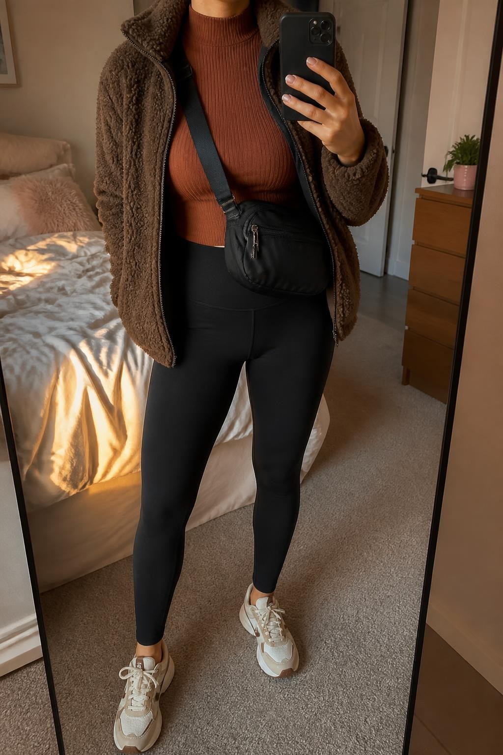 mirror selfie of a woman wearing an open chocolate brown sherpa fleece jacket over a rust fitted ribbed mock-neck top, black high-waisted ponte leggings, cream and taupe supportive sneakers, and a black nylon crossbody bag resting near the waist, clearly showing the exact clothing items, colors, fabrics, sneakers, and bag listed in the Outfit Pieces section, holding a phone in front of a mirror, cropped at NECK so the face does not show, relaxed sporty selfie pose in a feminine-themed bedroom, evening golden hour light, with soft pillows on an unmade bed and a small plant on a dresser, realistic sherpa fleece, ribbed knit, ponte, rubber, and nylon textures, latest iPhone photo quality
