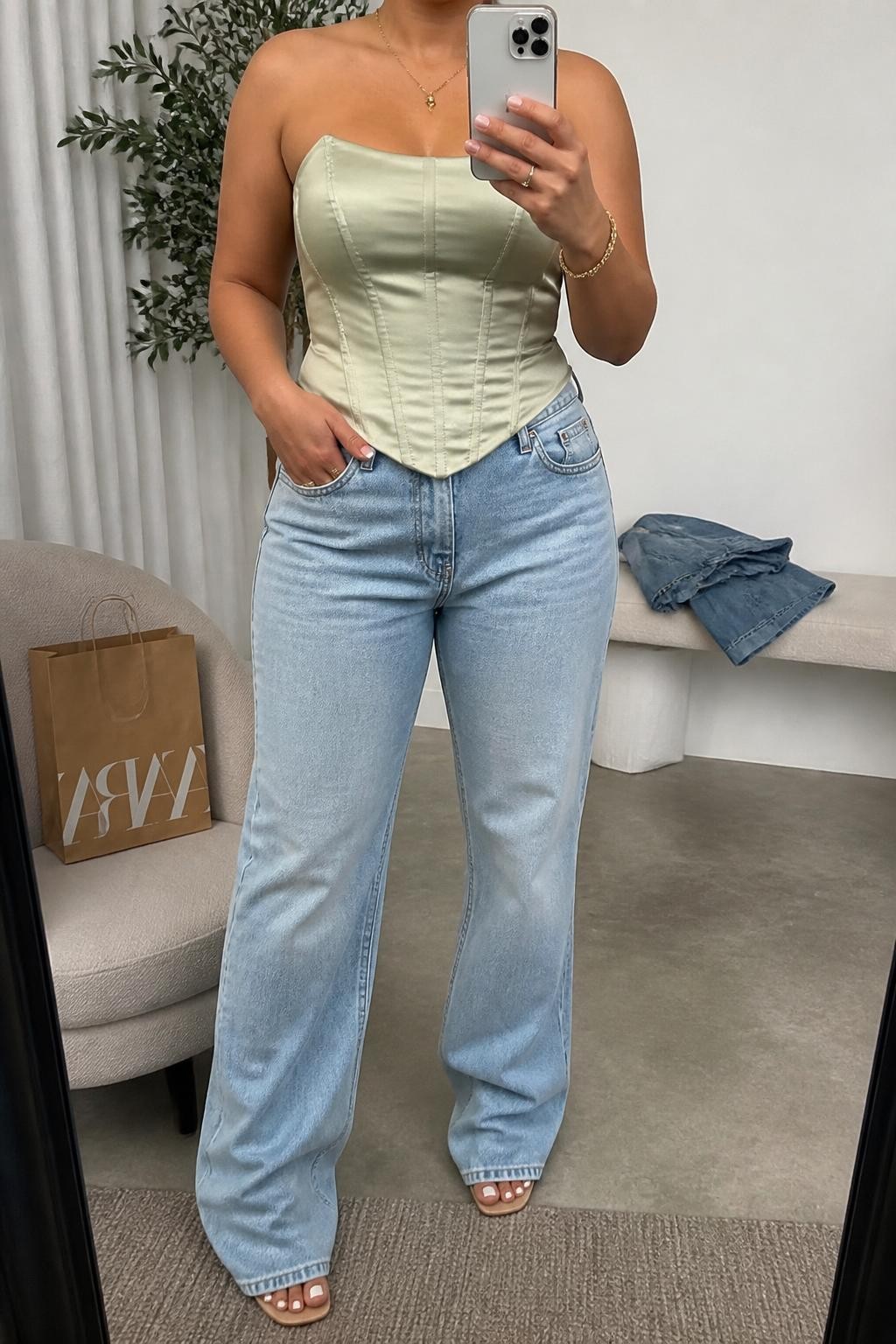 Mirror selfie of a plus size woman wearing Light-Wash Boyfriend Jeans & Satin Corset Top, clearly showing the exact outfit pieces, fabrics, colors, and accessories listed, holding a phone in front of a mirror, cropped at the neck so the face is hidden, natural casual selfie pose, in a fashion studio mirror corner with soft indoor lighting, a tote-sized shopping bag beside a chair and folded denim jeans on a bench, bright realistic iPhone appearance, latest iPhone photo quality.