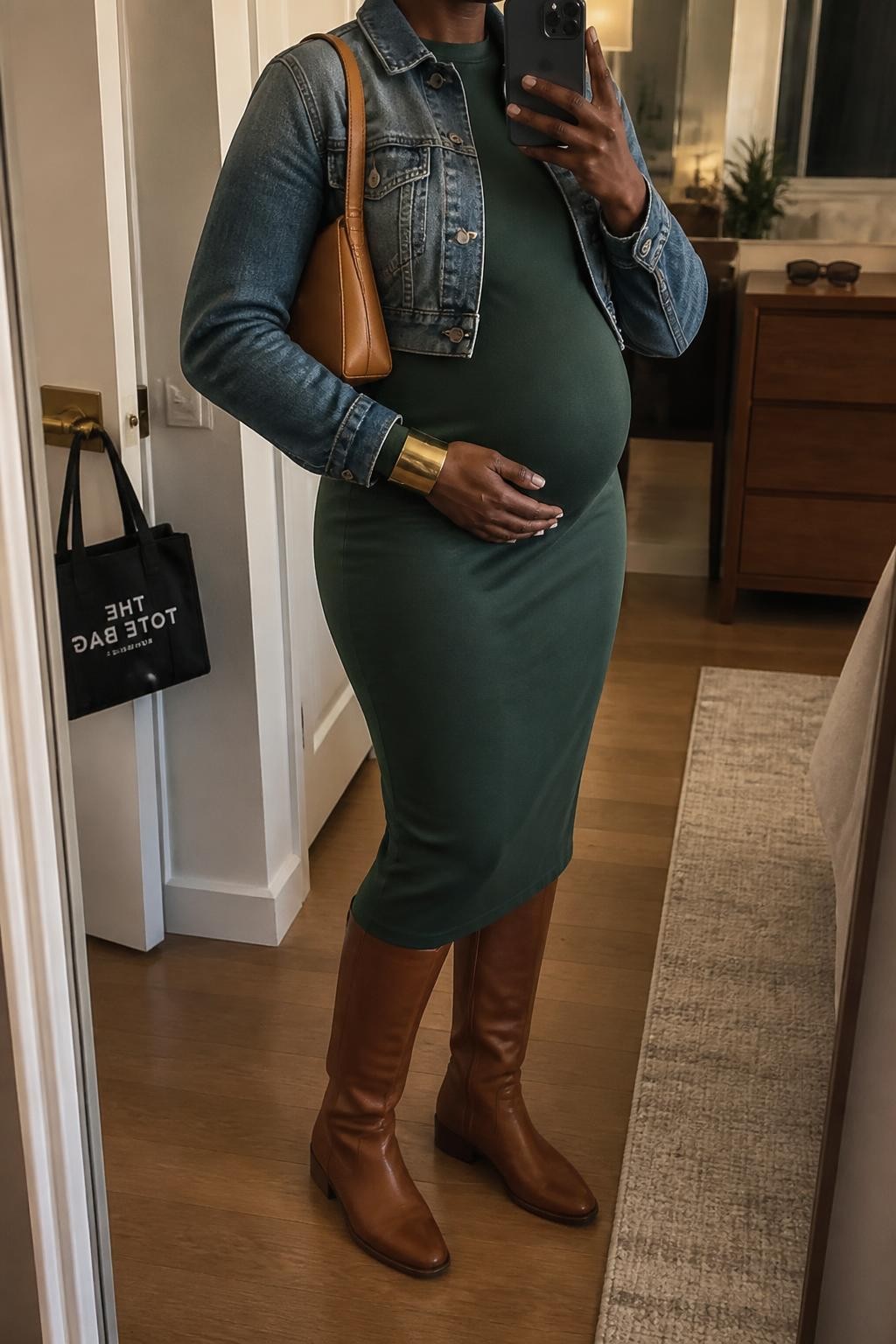 mirror selfie of a Black pregnant woman wearing a forest green long-sleeve maternity midi dress fitted comfortably around her visible baby bump, an open medium-wash cropped denim jacket ending above the bump, brown leather flat knee-high boots, a tan structured shoulder bag, and a wide brushed gold bracelet, clearly showing the exact clothing items, colors, fabrics, boots, bag, and bracelet listed in the Outfit Pieces section, holding a phone in front of a mirror, cropped at NECK so the face does not show, relaxed confident maternity selfie pose in a stylish city apartment, warm moody lighting, with a handbag hanging on a door handle and sunglasses placed on a dresser, realistic stretch fabric, denim, and leather textures, latest iPhone photo quality