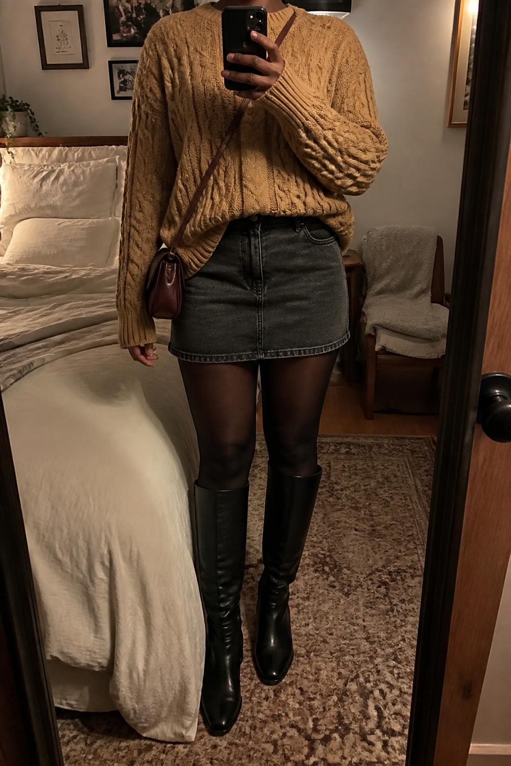 Mirror selfie of a Black woman wearing a faded black denim mini skirt, oversized camel cable-knit sweater, opaque black tights, black leather knee-high boots, and an oxblood crossbody bag, clearly showing the exact clothing items, colors, fabrics, and accessories listed, holding a phone in front of a cozy bedroom mirror setup, cropped at the neck so the face does not show, relaxed casual selfie pose, warm moody lighting, soft pillows on an unmade bed and a folded blanket on a chair, realistic lived-in setting, latest iPhone photo quality.