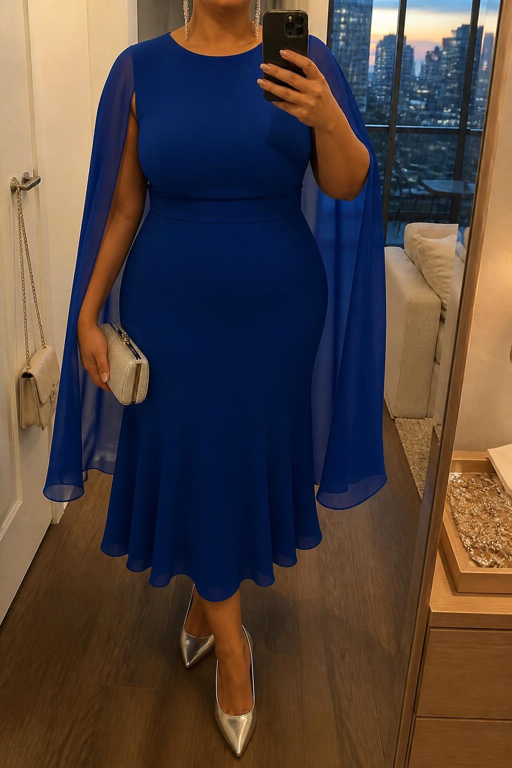 Mirror selfie of a plus size woman wearing a sapphire blue midi dress with a fitted sleeveless bodice, a softly defined waist, and a lightweight chiffon cape attached at the shoulders, metallic silver pointed heels, a silver hard-case clutch, and long silver linear earrings, clearly showing the exact dress shape, blue color, chiffon cape, footwear, and accessories listed, holding a phone in front of a mirror, cropped at the neck so the face does not show, relaxed dramatic selfie pose, in a stylish city apartment with evening golden-hour lighting, a handbag hanging on a door handle and a jewelry tray on the dresser, realistic formal wedding reception appearance, latest iPhone photo quality.