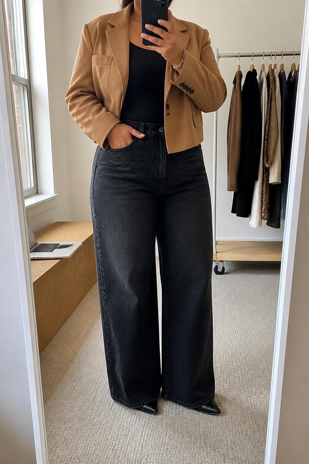 Mirror selfie of a plus size woman wearing Black Wide-Leg Jeans & Camel Cropped Blazer, clearly showing the same outfit pieces, colors, textures, and accessories listed, holding a phone in front of a mirror, cropped at the neck so her face is not visible, relaxed casual stance, in a minimal dressing room with natural window light, a laptop resting on a bench and a clothing rack with garments behind her, realistic everyday mirror selfie, latest iPhone photo quality.