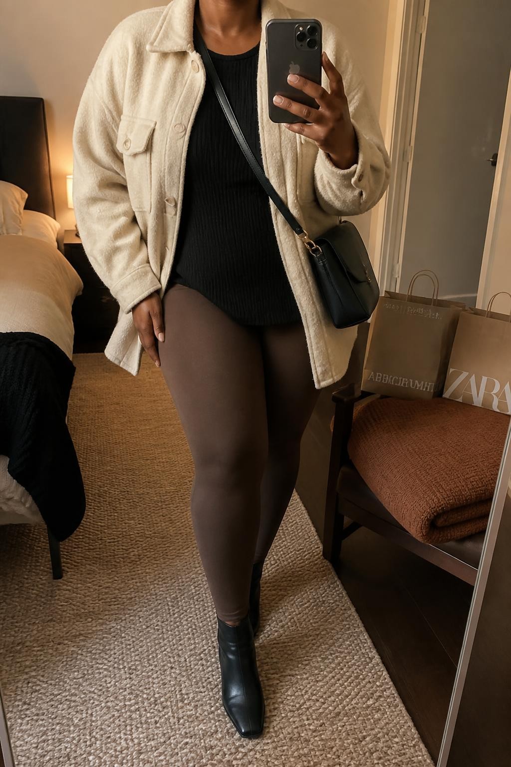 mirror selfie of a Black curvy woman wearing an open oatmeal brushed wool-blend shacket over a black longline ribbed knit top falling smoothly over chocolate high-waisted ponte leggings, black leather ankle boots, and a black structured crossbody bag, clearly showing the exact clothing items, colors, fabrics, boots, and bag listed in the Outfit Pieces section, holding a phone in front of a mirror, cropped at NECK so the face does not show, relaxed casual selfie pose in a cozy bedroom mirror setup, warm moody lighting, with a folded blanket on a chair and shopping bags beside it, realistic brushed wool, ribbed knit, ponte, and leather textures, natural unstaged mirror photo, latest iPhone photo quality