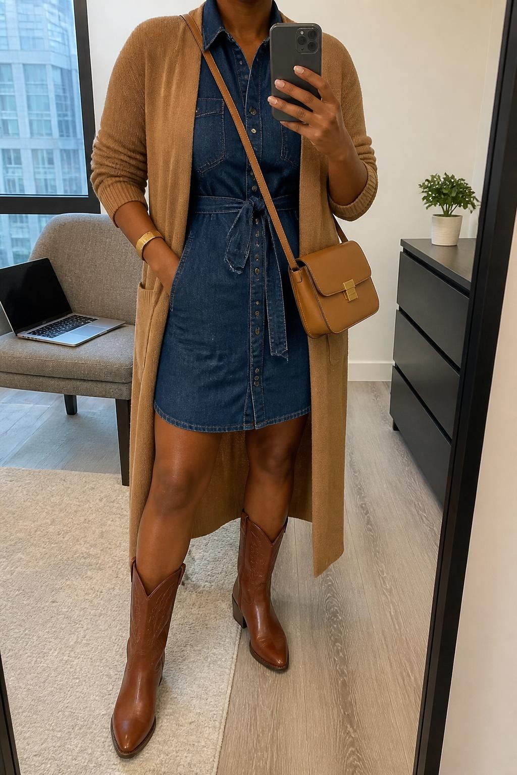 Image Prompt mirror selfie of a Black woman wearing a dark-wash belted denim shirt dress tied at the natural waist, a fully open camel longline cardigan, chestnut leather cowboy boots, a tan structured crossbody bag, and a slim gold cuff bracelet, clearly showing the exact clothing items, colors, fabrics, boots, bag, and bracelet listed in the Outfit Pieces section, holding a phone in front of a mirror, cropped at NECK so the face does not show, relaxed confident selfie pose in a stylish city apartment, soft indoor lighting, with a laptop resting on a chair and a small plant on a dresser, realistic denim, cardigan knit, and leather textures, latest iPhone photo quality