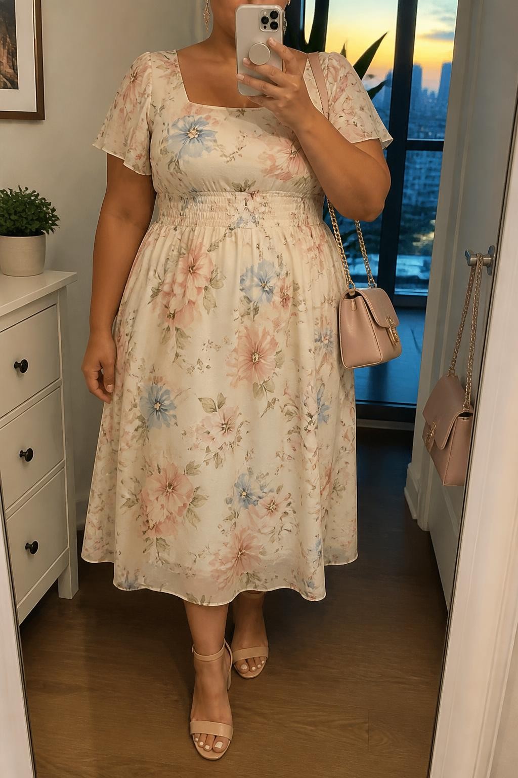 Mirror selfie of a plus size woman wearing a cream chiffon midi dress with blush pink, pale blue, and sage floral prints, short flutter sleeves, a square neckline, and a softly smocked waist, nude block-heel sandals, a blush pink shoulder bag, and delicate gold drop earrings, clearly showing the exact floral dress, print colors, smocked waist, footwear, and accessories listed, holding a phone in front of a mirror, cropped at the neck so her face is hidden, relaxed graceful selfie stance, in a stylish city apartment with evening golden-hour lighting, a small plant on a dresser and a handbag hanging on a door handle, realistic baby shower appearance, latest iPhone photo quality.