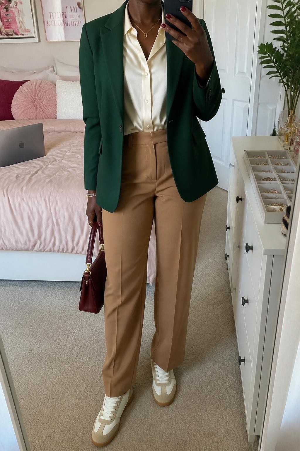mirror selfie of a Black woman wearing an open forest green tailored blazer over an ivory satin button-front blouse tucked into camel high-waisted straight-leg trousers, cream retro-style sneakers with tan suede panels, and an oxblood structured handbag, clearly showing the exact clothing items, colors, fabrics, sneakers, and bag listed in the Outfit Pieces section, holding a phone in front of a mirror, cropped at NECK so the face does not show, relaxed professional selfie pose in a feminine-themed bedroom, soft indoor lighting, with a laptop on the bed and a jewelry tray on a dresser, realistic tailoring, satin, trouser fabric, leather, and suede textures, latest iPhone photo quality