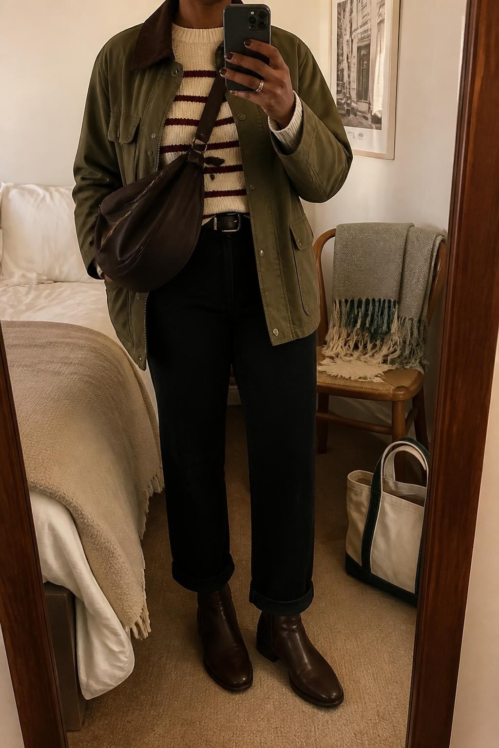mirror selfie of a Black woman wearing an olive cotton-look barn jacket with a brown corduroy collar partly open over a cream and burgundy striped knit sweater, black high-rise straight-leg jeans cuffed once above dark brown flat ankle boots, and a chocolate nylon crossbody bag, clearly showing the exact clothing items, colors, striped pattern, fabrics, boots, and bag listed in the Outfit Pieces section, holding a phone in front of a mirror, cropped at NECK so the face does not show, relaxed casual selfie pose in a cozy bedroom mirror setup, warm moody lighting, with a folded blanket on a chair and a tote bag resting on the floor, realistic cotton-look fabric, corduroy, knit, denim, leather-look, and nylon textures, latest iPhone photo quality
