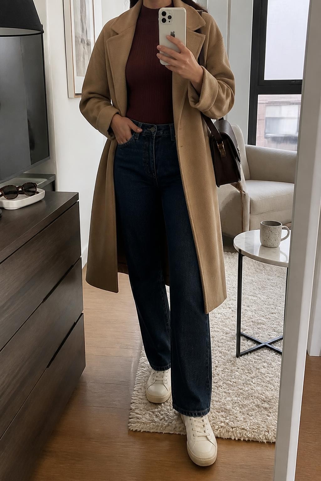 mirror selfie of a woman wearing an open camel knee-length wool-blend coat over a burgundy fitted ribbed mock-neck sweater, dark-wash high-rise straight-leg jeans, cream leather sneakers, and a chocolate structured shoulder bag, clearly showing the exact clothing items, colors, fabrics, sneakers, and bag listed in the Outfit Pieces section, holding a phone in front of a full-length mirror, cropped at NECK so the face does not show, relaxed confident selfie pose in a stylish city apartment, natural window light, with sunglasses placed on a dresser and a coffee mug on a side table, realistic wool, ribbed knit, denim, and leather textures, latest iPhone photo quality
