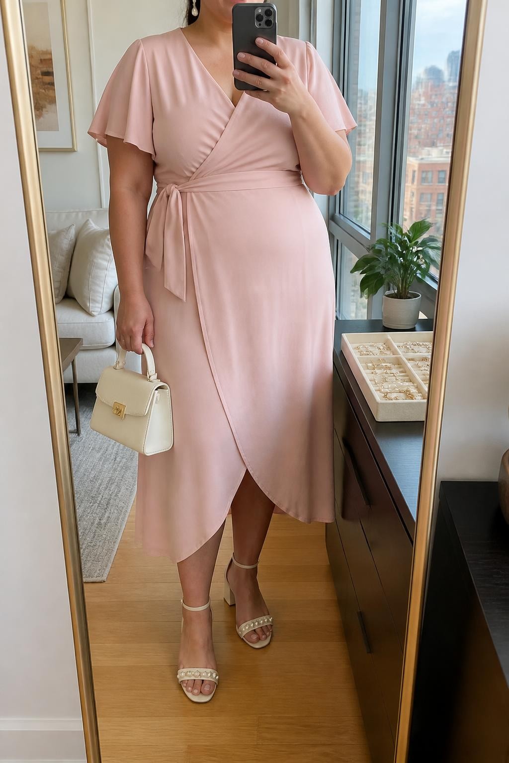 Mirror selfie of a plus size woman wearing a blush pink crepe wrap midi dress with short flutter sleeves, a modest V-neckline, and a matching waist tie, cream block-heel sandals with small pearl straps, a structured cream mini handbag, and pearl drop earrings, clearly showing the exact clothing items, blush crepe, wrap shape, cream footwear, pearl details, and accessories listed, holding a phone in front of a mirror, cropped at the neck so the face does not show, relaxed feminine selfie pose, in a stylish city apartment with natural window light, a jewelry tray on the dresser and a small plant nearby, natural unstaged baby shower appearance, latest iPhone photo quality.