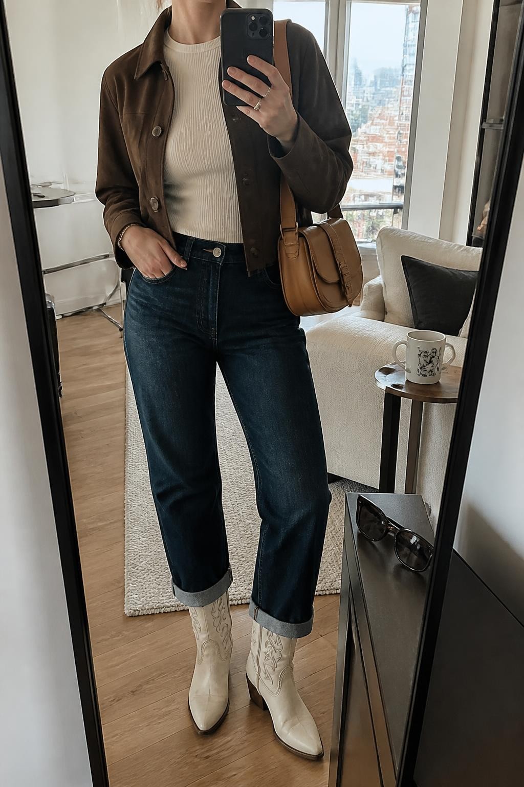 mirror selfie of a woman wearing a chocolate brown cropped suede jacket over an ivory fitted ribbed crewneck top, dark-wash straight-leg jeans cuffed once above cream leather cowboy boots with brown stitching, and a tan leather saddle bag, clearly showing the exact clothing items, colors, fabrics, boots, stitching, and bag listed in the Outfit Pieces section, holding a phone in front of a full-length mirror, cropped at NECK so the face does not show, relaxed casual selfie pose in a stylish city apartment, natural window light, with sunglasses placed on a dresser and a coffee mug on a side table, realistic suede, ribbed knit, denim, and leather textures, latest iPhone photo quality
