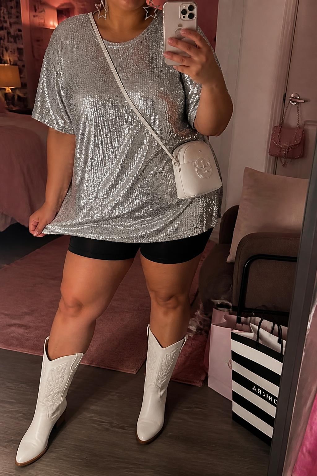 Mirror selfie of a plus size woman wearing a loose silver sequin T-shirt mini dress with short sleeves, fitted black anti-chafe shorts underneath, white mid-calf cowboy boots, a small white leather crossbody bag, and silver star-shaped earrings, clearly showing the exact clothing items, silver sequins, black shorts, white boots, colors, and accessories listed, holding a phone in front of a mirror, cropped at the neck so the face does not show, relaxed playful selfie pose, in a feminine-themed bedroom with warm moody lighting, shopping bags beside a chair and a handbag hanging on a door handle, natural evening festival appearance, latest iPhone photo quality.