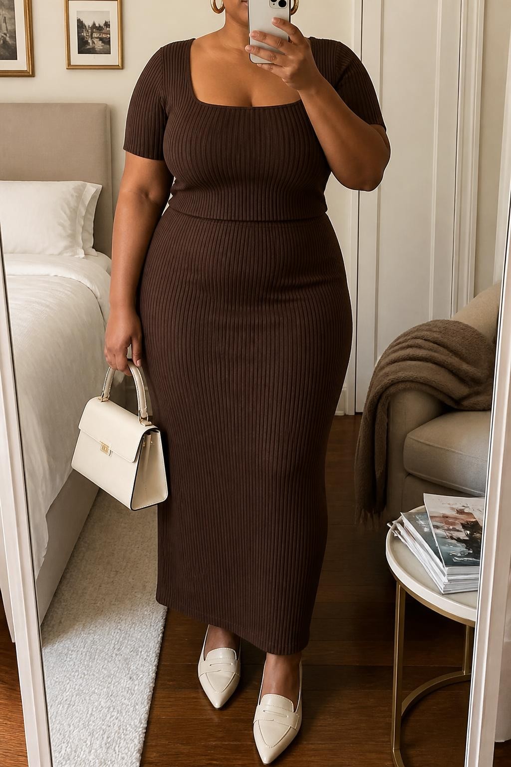 Mirror selfie of a plus size woman wearing a chocolate brown ribbed short-sleeve top with a square neckline, a matching chocolate brown high-waisted knit midi skirt, cream pointed leather loafers, a structured cream top-handle bag, and chunky gold hoop earrings, clearly showing the exact matching set, chocolate ribbed texture, cream footwear, and accessories listed, holding a phone in front of a mirror, cropped at the neck so her face does not show, relaxed sophisticated selfie stance, in a cozy bedroom mirror setup with soft indoor lighting, a folded blanket on a chair and a stack of fashion magazines on a side table, realistic cooler-weather brunch outfit, latest iPhone photo quality.