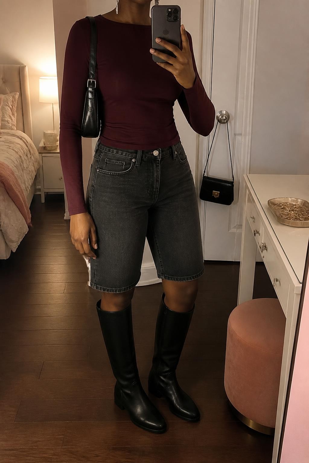 mirror selfie of a Black woman wearing faded charcoal gray denim jorts with a clean knee-length cut, a fitted burgundy long-sleeve stretch top, black knee-high leather boots with a low heel, a black mini shoulder bag and slim silver drop earrings, wearing exactly the listed outfit without additional garments or accessories, holding a phone in front of a mirror, cropped at NECK so the face does not show, relaxed casual selfie pose, in a feminine-themed bedroom, warm moody lighting, handbag hanging on a door handle and a jewelry tray on the vanity, realistic transitional styling, latest iPhone photo quality.
