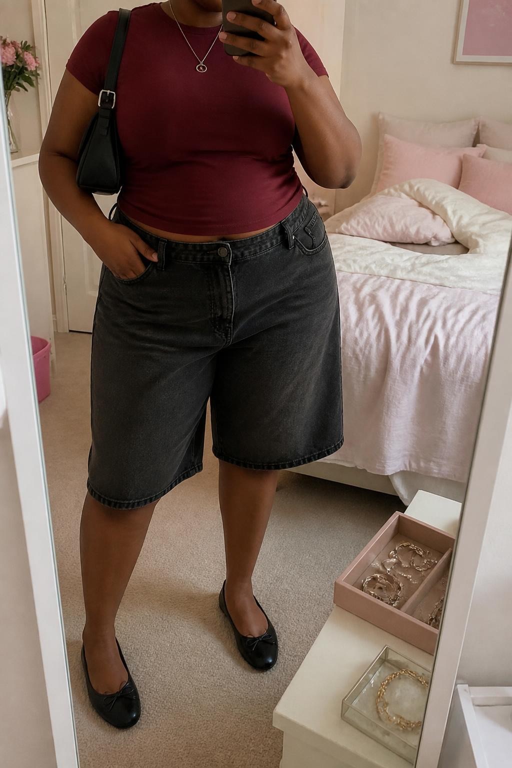 mirror selfie of a plus size Black woman wearing baggy washed black denim jorts with a loose knee-length cut, a fitted burgundy cotton baby tee, black leather ballet flats, a compact black shoulder bag and a thin silver pendant necklace, wearing exactly the listed outfit with no added clothing pieces, holding a phone in front of a mirror, cropped at NECK so the face does not show, relaxed casual selfie pose, in a feminine-themed bedroom, soft indoor lighting, soft pillows on an unmade bed and a jewelry tray on the vanity, believable everyday styling, latest iPhone photo quality.