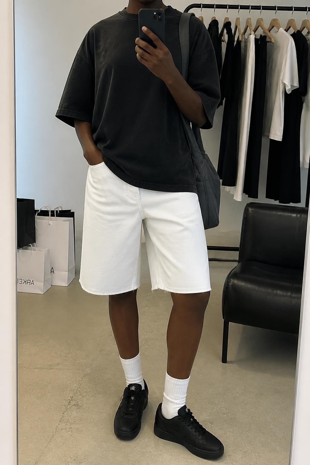 mirror selfie of a Black woman wearing white Bermuda denim jorts with a clean straight fit, an oversized washed black heavyweight cotton T-shirt with no graphics, black low-profile sneakers, white ribbed crew socks and a charcoal nylon shoulder bag, wearing exactly the listed outfit with no additional pieces, holding a phone in front of a mirror, cropped at NECK so the face does not show, relaxed street-style selfie pose, in a fashion studio mirror corner, soft indoor lighting, clothing rack with garments behind and shopping bags beside a chair, natural casual latest iPhone photo quality.