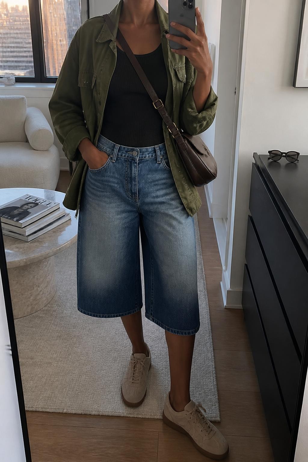 mirror selfie of a Black woman wearing washed blue long denim jorts with a loose below-the-knee shape, an open olive cotton utility jacket over a fitted black ribbed tank, beige suede sneakers and a dark brown leather crossbody bag, wearing exactly the listed clothing and accessories with nothing additional, holding a phone in front of a mirror, cropped at NECK so the face does not show, relaxed casual selfie pose, in a stylish city apartment, evening golden hour, stack of fashion magazines on a side table and sunglasses placed on a dresser, realistic transitional styling, latest iPhone photo quality.